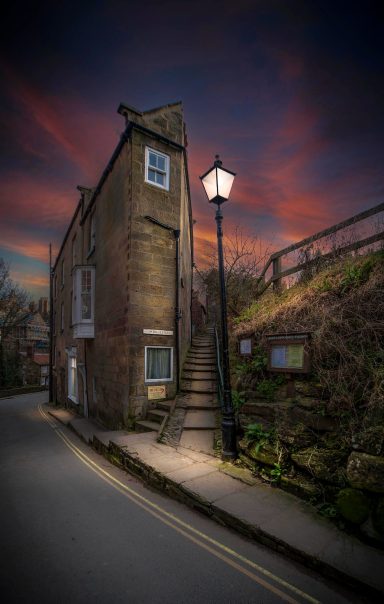 Jim Bell's stile at dusk Stone building with a staircase and a lamp post, silhouetted against a dramatic sunset sky.