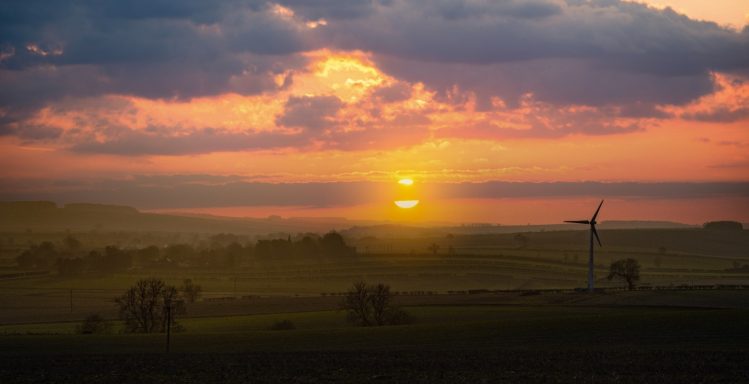 Sunset over a landscape with a wind turbine and distant trees against a cloudy sky.