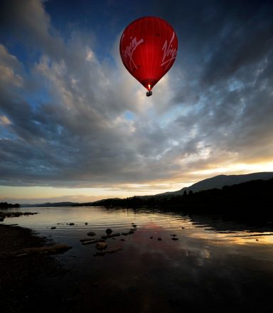 Red hot air balloon over a calm lake at sunset, reflecting the sky and surrounding landscape.