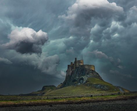 Lindisfarne Castle Dramatic clouds loom over a castle on a rocky hill, surrounded by green landscape.