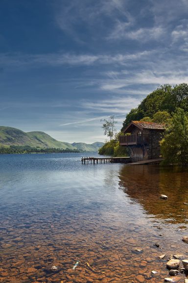 The Duke of Portland Boathouse Ullswater, Scenic lake view with a rustic boathouse and green hills under a blue sky.